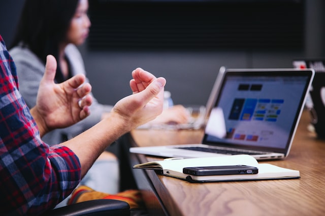 photo of person moving their hands while talking in front of a computer with another person listening