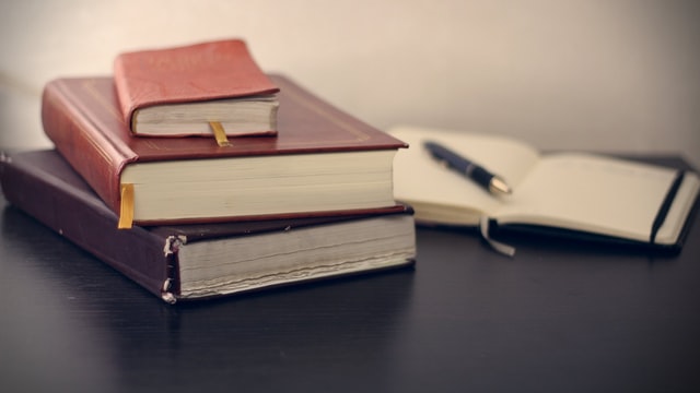 a stack of books next to an open book to blank pages with a pen sitting on it