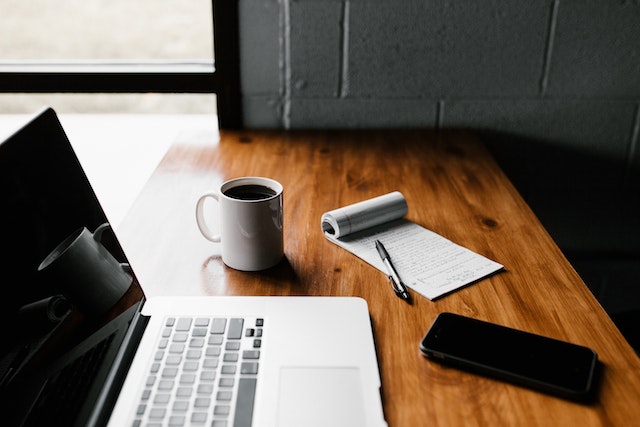 photo of a laptop, mobile phone and notepad and pen on a wooden desk