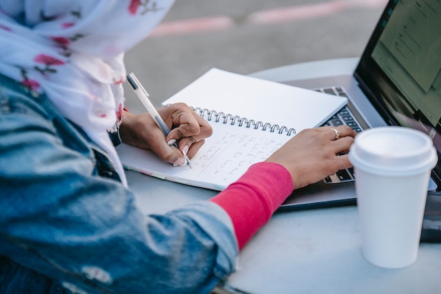 woman with a headscarf writing notes in notebook while using laptop