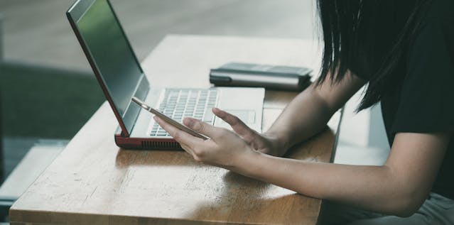 close up photo of a person holding a mobile phone with a laptop on the desk