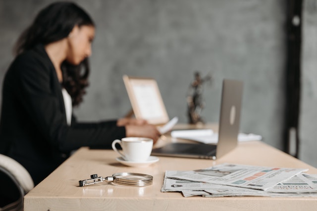 photo slightly out of focus of a woman working at a desk with laptop and lady justice in the background, with a newspaper and magnifying glass